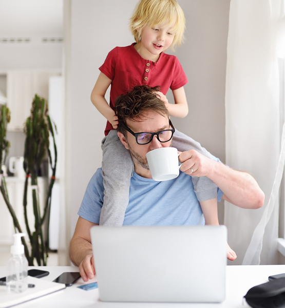 A man drinking his coffee sitting down at a home desk wearing a blue top whilst his child wearing a red top is sitting on his shoulders, the background is a well lit white office type room
