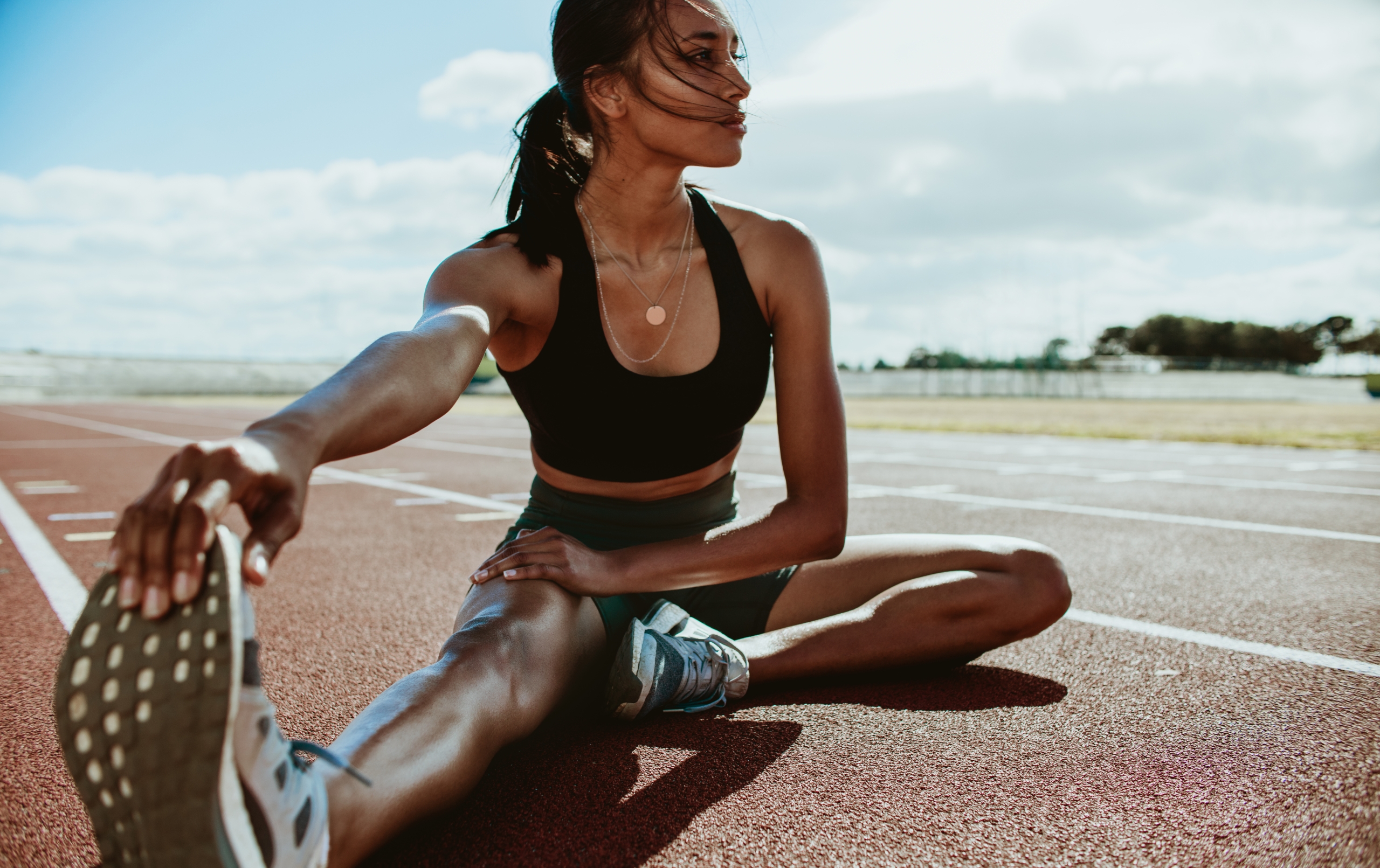 a girl in gym wear stretching on the floor laying in the middle of a running track in a sunny background.