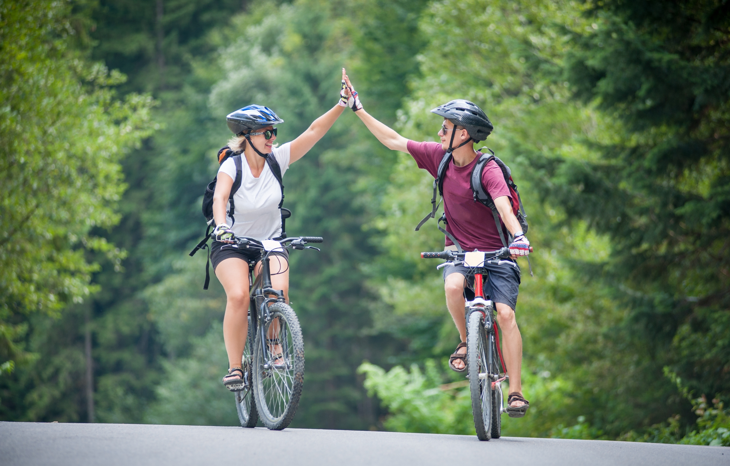 a male and a female on seperate bicycles slapping their hands together both smiling at eachother riding down a road with forest surroundings